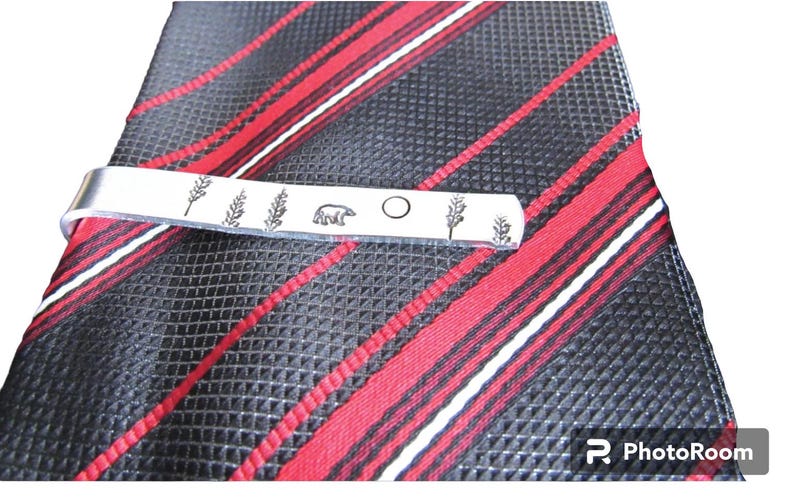 May include: Silver tie clip with a bear, a circle, and two sprigs of wheat engraved on it. The tie clip is on a red and black striped tie.