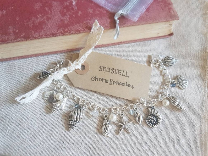 May include: A silver charm bracelet with a seashell theme. The bracelet features a variety of charms, including seashells, starfish, and pearls. The bracelet is on a brown fabric background with a tag that reads "Seashell Charm Bracelet".