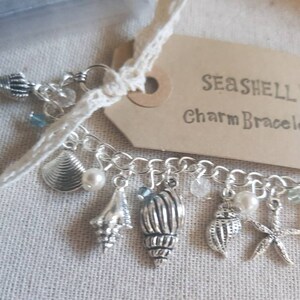 May include: A silver charm bracelet with a seashell theme. The bracelet features a variety of charms, including seashells, starfish, and beads. The tag attached to the bracelet reads "Seashell Charm Bracelet".
