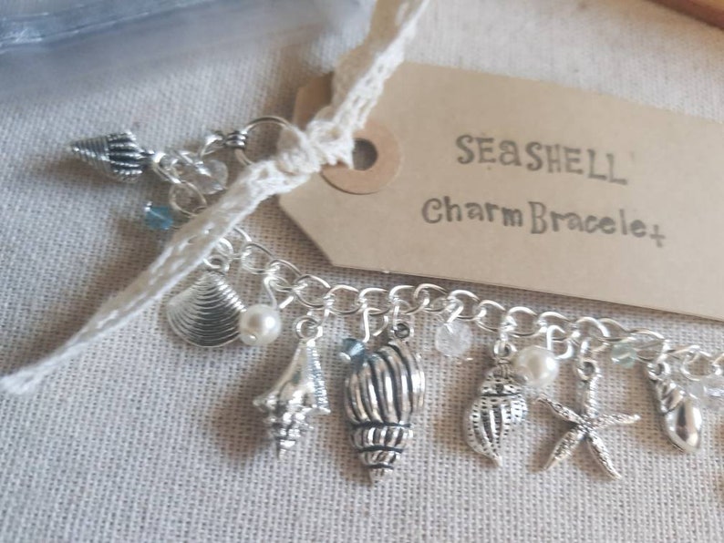 May include: A silver charm bracelet with a seashell theme. The bracelet features a variety of charms, including seashells, starfish, and beads. The tag attached to the bracelet reads "Seashell Charm Bracelet".
