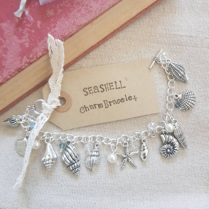 May include: A silver charm bracelet with a seashell theme. The bracelet features a variety of charms, including seashells, starfish, and pearls. The tag attached to the bracelet reads "Seashell Charm Bracelet".
