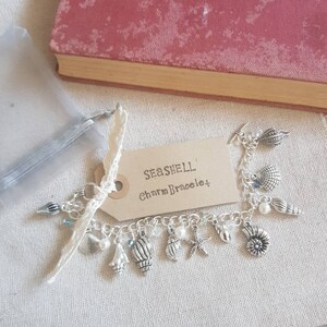 May include: A silver charm bracelet with a variety of seashell charms, including starfish, seashells, and pearls. The bracelet is on a brown tag that says "Seashell Charm Bracelet".