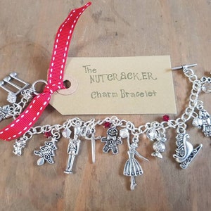 May include: A silver charm bracelet with a red and white ribbon tied around it. The bracelet has charms of a music note, a pair of ballet shoes, a Christmas tree, a nutcracker soldier, a sword, a gingerbread man, a bell, a candy cane, a sleigh, and a snowflake. The tag attached to the ribbon reads "The Nutcracker Charm Bracelet".