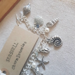 May include: A silver charm bracelet with a seashell theme. The bracelet features a variety of charms, including seashells, starfish, and pearls. The bracelet is labeled "Seashell Charm Bracelet".