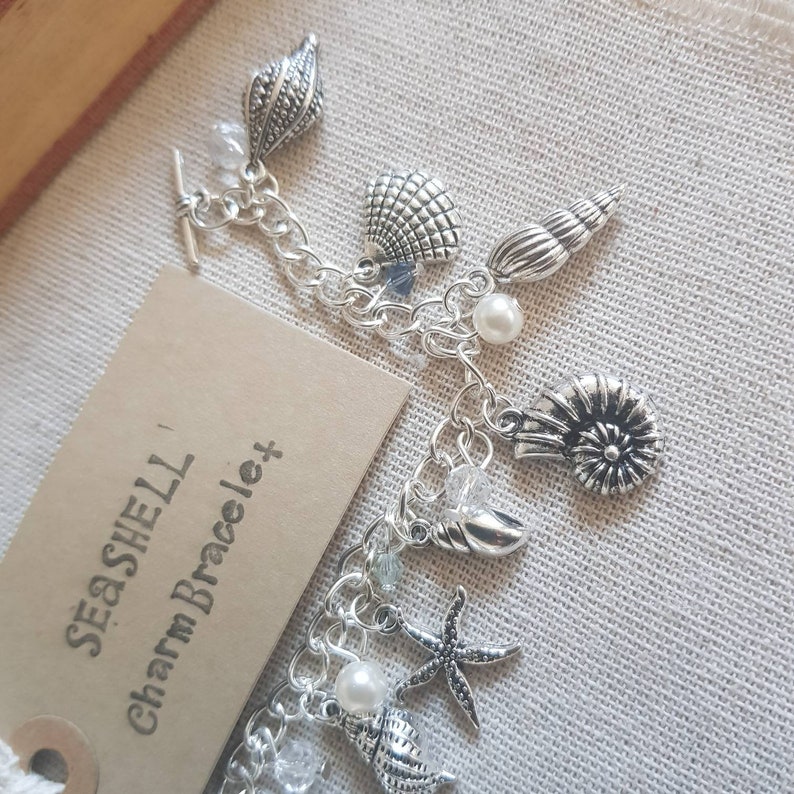 May include: A silver charm bracelet with a seashell theme. The bracelet features a variety of charms, including seashells, starfish, and pearls. The bracelet is labeled "Seashell Charm Bracelet".
