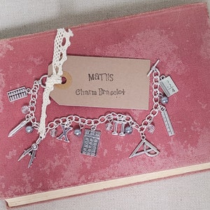 May include: A silver charm bracelet with math-themed charms, including an abacus, pencil, compass, calculator, and ruler. The bracelet is attached to a tag that reads "MATHS Charm Bracelet" with a white lace ribbon. The bracelet rests on a vintage, dusty rose book.
