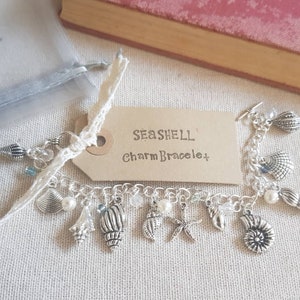 May include: A silver charm bracelet with a seashell theme. The bracelet has a variety of charms, including seashells, starfish, and pearls. The bracelet is on a white background with a brown tag that says "Seashell Charm Bracelet".