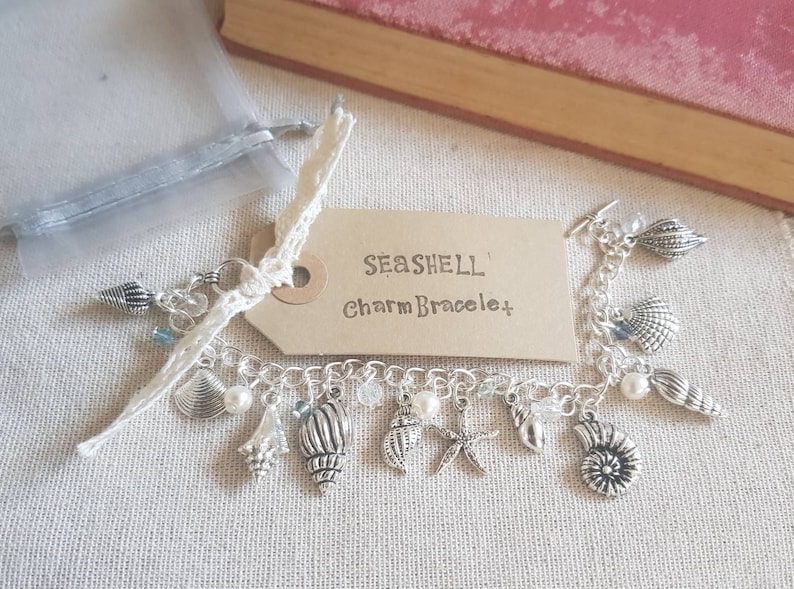 May include: A silver charm bracelet with a seashell theme. The bracelet has a variety of charms, including seashells, starfish, and pearls. The bracelet is on a white background with a brown tag that says "Seashell Charm Bracelet".