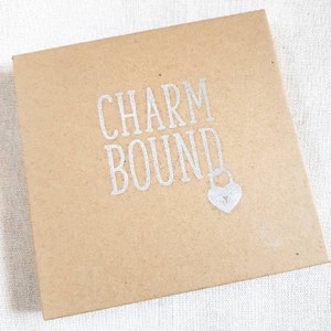 May include: A brown cardboard box with the text "CHARM BOUND" printed in silver lettering. A small silver heart-shaped padlock is below the text.