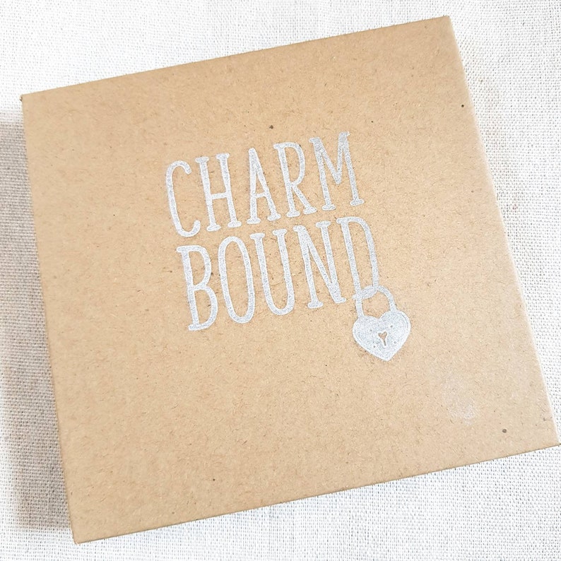 May include: A brown cardboard box with the text "CHARM BOUND" printed in silver lettering. A small silver heart-shaped padlock is below the text.