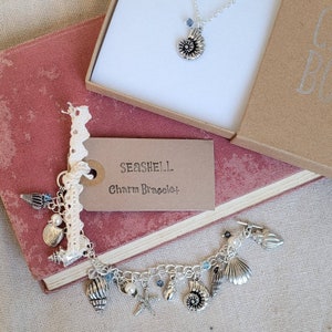 May include: A silver charm bracelet with a seashell theme. The bracelet features a variety of charms, including seashells, starfish, and pearls. The bracelet is displayed on a book with a tag that reads "Seashell Charm Bracelet".