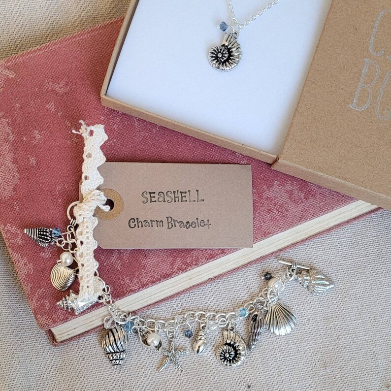 May include: A silver charm bracelet with a seashell theme. The bracelet features a variety of charms, including seashells, starfish, and pearls. The bracelet is displayed on a book with a tag that reads "Seashell Charm Bracelet".