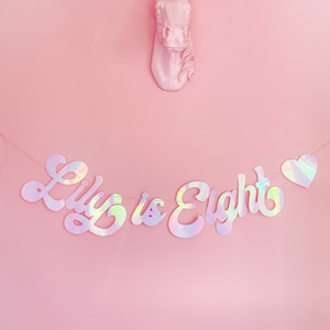 May include: A pink unicorn head with a gold horn hangs on a pink wall with a iridescent banner that reads "Lily is Eight".