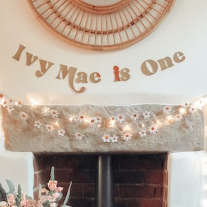 May include: A decorative wall display featuring a woven rattan mirror, a banner that reads "Ivy Mae is One", and a daisy chain light garland. The banner letters are in a light brown color, with the word "is" in red.