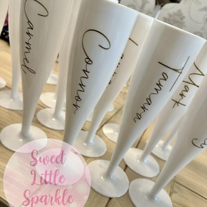 May include: A group of white champagne flutes with gold calligraphy names. The flutes are displayed on a light-coloured wooden surface. The text "Sweet Little Sparkle" is in a pink circle in the lower left corner.
