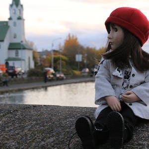 Pode incluir: Uma boneca usando um gorro vermelho e um casaco cinza est&aacute; sentada em uma borda de concreto. A boneca tem cabelo castanho longo e est&aacute; olhando para a esquerda. A boneca est&aacute; sentada em frente a um corpo de &aacute;gua e um pr&eacute;dio no fundo.