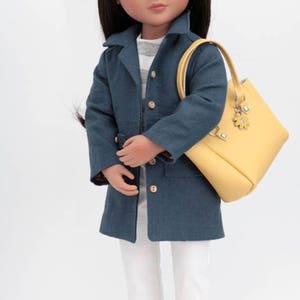 Pode incluir: Uma boneca vestindo um casaco azul, cal&ccedil;as brancas e sapatos brancos. A boneca est&aacute; segurando uma bolsa amarela com um chaveiro de flor.