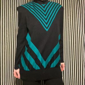 May include: A black long-sleeve mock neck top featuring a bold teal chevron pattern across the chest and lower torso. The garment has a solid black ribbed hem and solid black sleeves, creating a sharp contrast with the geometric teal stripes.