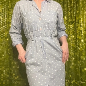 May include: A light blue button-down dress featuring a repeating white abstract pattern. The garment includes a collared neckline, long sleeves with cuffs, two chest pockets, a cinched waist with belt loops, and a straight skirt with a front slit.