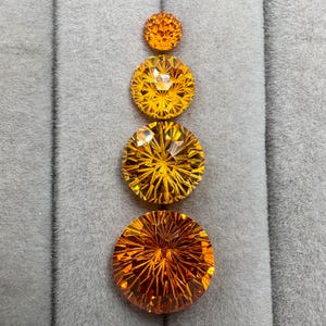 Pode incluir: Quatro pedras preciosas redondas e facetadas em vários tamanhos, dispostas verticalmente. As gemas são de uma cor laranja vibrante, com cortes internos intrincados que refletem a luz. As pedras preciosas são exibidas em uma superfície cinza texturizada.