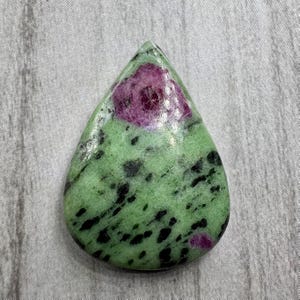 Puede incluir: Una gema en forma de lágrima con una base verde, motas negras y un centro magenta intenso. La superficie pulida refleja la luz, resaltando los patrones naturales y las texturas de la piedra.