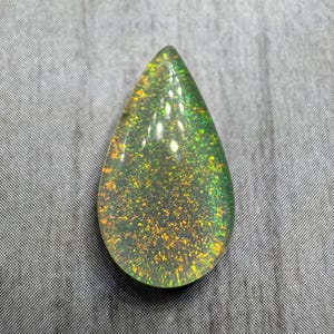 May include: A teardrop-shaped gemstone with a shimmering, iridescent effect. The gem features a clear base with embedded flecks of gold and green, creating a sparkling appearance. The stone is polished and smooth.