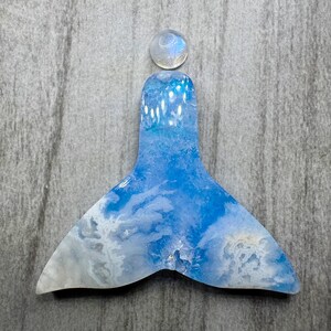 May include: A blue and white resin mermaid tail with a round, clear stone at the top. The tail has a cloudy, marbled design. The stone is a light, iridescent color. The background is a gray, textured surface.
