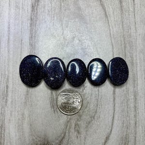 May include: Five oval-shaped, dark blue gemstones with sparkling inclusions are arranged in a row. A quarter coin is placed below the gemstones for size comparison. The stones have a polished, reflective surface.