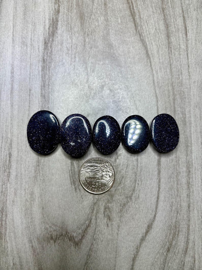 May include: Five oval-shaped, dark blue gemstones with sparkling inclusions are arranged in a row. A quarter coin is placed below the gemstones for size comparison. The stones have a polished, reflective surface.