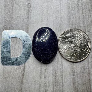 May include: A dark blue, oval-shaped gemstone with sparkling inclusions, next to a silver metal letter "D" and a United States quarter coin. The gemstone's surface reflects light, creating a starry effect. The coin is a 25-cent piece.