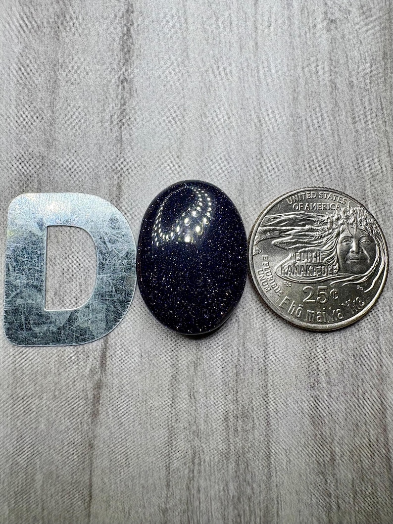 May include: A dark blue, oval-shaped gemstone with sparkling inclusions, next to a silver metal letter "D" and a United States quarter coin. The gemstone's surface reflects light, creating a starry effect. The coin is a 25-cent piece.