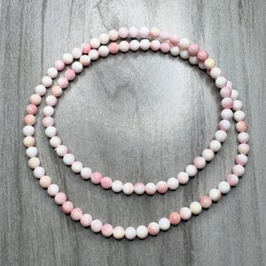 May include: A long strand of smooth, spherical beads in varying shades of soft pink and white. The beads are arranged in a circular pattern, showcasing a consistent size and polished finish throughout the entire length of the beaded necklace strand.