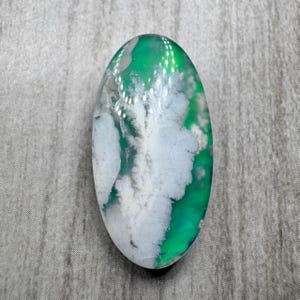 May include: An oval-shaped gemstone with a vibrant green and white pattern. The stone's surface features a mix of translucent green and opaque white, creating a cloudy, natural design. The gemstone is set against a gray background.