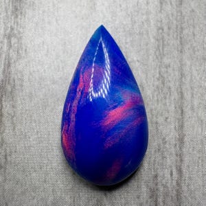 May include: A teardrop-shaped gemstone with a vibrant blue base color, swirled with streaks of pink and lighter blue. The polished surface reflects light, highlighting the unique patterns within the stone. The gemstone is set against a gray textured background.