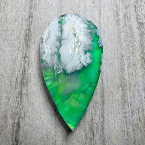 May include: A teardrop-shaped gemstone with a vibrant green base, transitioning to white, cloud-like patterns at the top. The stone has a polished, glossy finish, showcasing the interplay of colors and textures. The gemstone is a unique piece of jewelry.