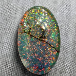 May include: An oval-shaped cabochon gemstone featuring a dark base with vibrant, multi-colored iridescent flecks in shades of red, green, and orange. The surface displays a distinct, dark, jagged crack running diagonally across the upper section.
