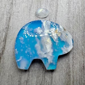 May include: A blue and white elephant-shaped stone carving with a small, oval, iridescent stone above it. The elephant carving has a glossy finish and a cloudy, ocean-like pattern. The background is a gray, textured surface.