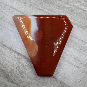 May include: A red and white agate cabochon with a triangular shape. The agate has a white band with brown and orange inclusions.