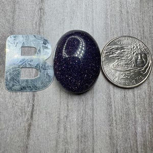 May include: A polished, oval-shaped blue sandstone gemstone with sparkling inclusions. Next to the gemstone is a silver metal letter "B" and a United States quarter coin.