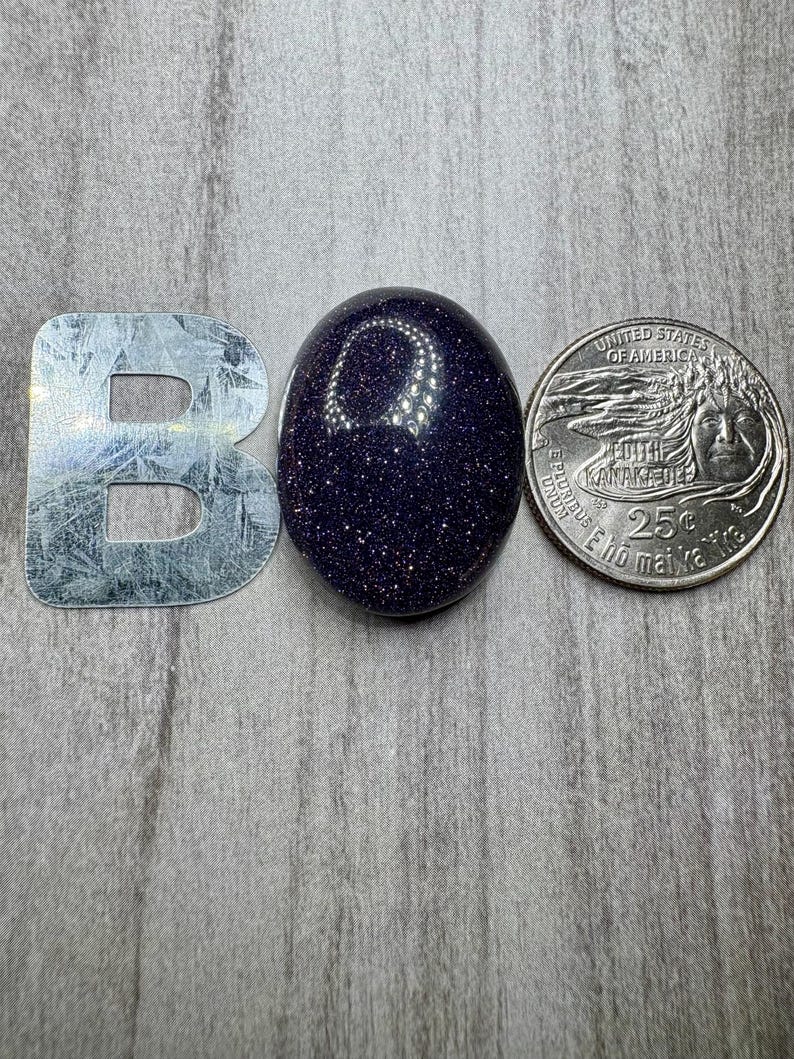 May include: A polished, oval-shaped blue sandstone gemstone with sparkling inclusions. Next to the gemstone is a silver metal letter "B" and a United States quarter coin.