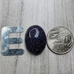 May include: A dark blue, oval-shaped gemstone with sparkling inclusions, next to a metal letter "E" and a U.S. quarter coin. The gemstone's surface reflects light, creating a starry effect. The coin is a quarter.