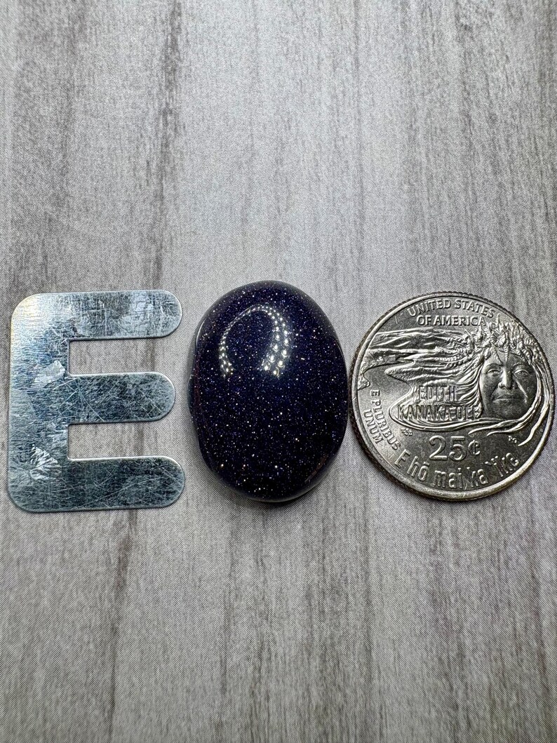 May include: A dark blue, oval-shaped gemstone with sparkling inclusions, next to a metal letter "E" and a U.S. quarter coin. The gemstone's surface reflects light, creating a starry effect. The coin is a quarter.