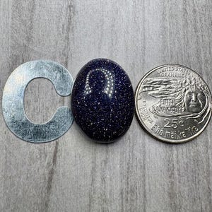 May include: A dark blue, oval-shaped gemstone with sparkling inclusions, next to a silver metal letter "C" and a United States quarter. The gemstone's surface reflects light, creating a starry effect. The quarter is a 25-cent coin.