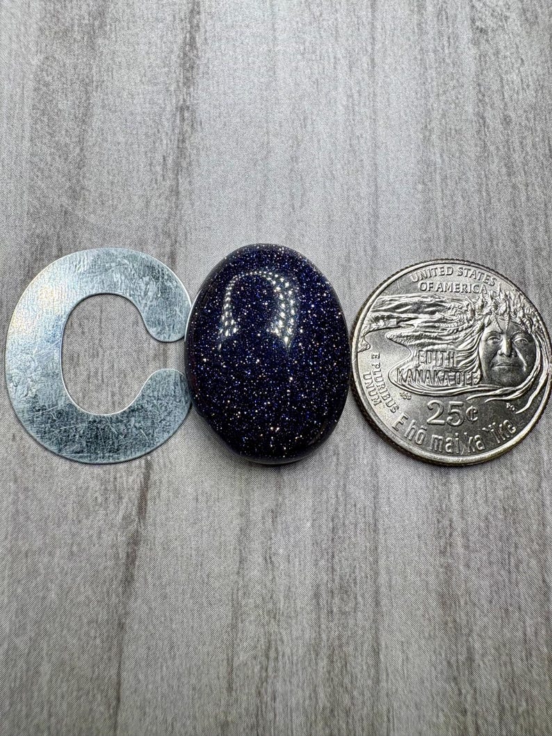 May include: A dark blue, oval-shaped gemstone with sparkling inclusions, next to a silver metal letter "C" and a United States quarter. The gemstone's surface reflects light, creating a starry effect. The quarter is a 25-cent coin.