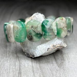 Puede incluir: Una pulsera elástica compuesta por cuentas lisas, pulidas y de forma ovalada en varios tonos de verde translúcido, blanco opaco y gris moteado. Las cuentas presentan inclusiones naturales y variaciones de color con aspecto jaspeado.