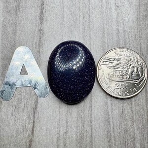 May include: A dark blue, oval-shaped gemstone with sparkling inclusions is displayed next to a silver letter "A" and a U.S. quarter. The gemstone's surface reflects light, and the quarter shows the profile of a woman.