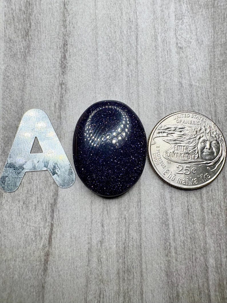 May include: A dark blue, oval-shaped gemstone with sparkling inclusions is displayed next to a silver letter "A" and a U.S. quarter. The gemstone's surface reflects light, and the quarter shows the profile of a woman.