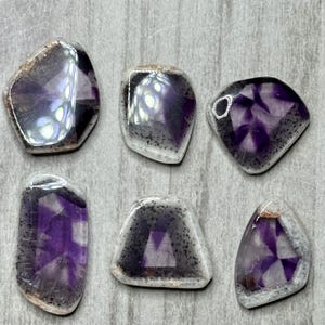 May include: Six polished gemstones with a mix of purple, gray, and clear colors. The stones have irregular shapes and a speckled pattern around the edges. The purple hues vary in intensity, creating a visually appealing display.