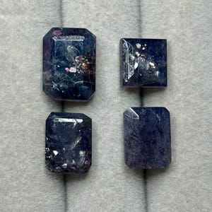 Puede incluir: Cuatro gemas facetadas de color azul oscuro, de forma rectangular o cuadrada. Las gemas tienen inclusiones internas y una superficie pulida. Las piedras preciosas están dispuestas sobre una superficie gris neutra.