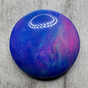 May include: A round, polished stone with a vibrant color palette. The stone features a deep blue base with swirling accents of pink, purple, and teal. A bright, curved highlight is visible on the top surface, adding dimension to the design.
